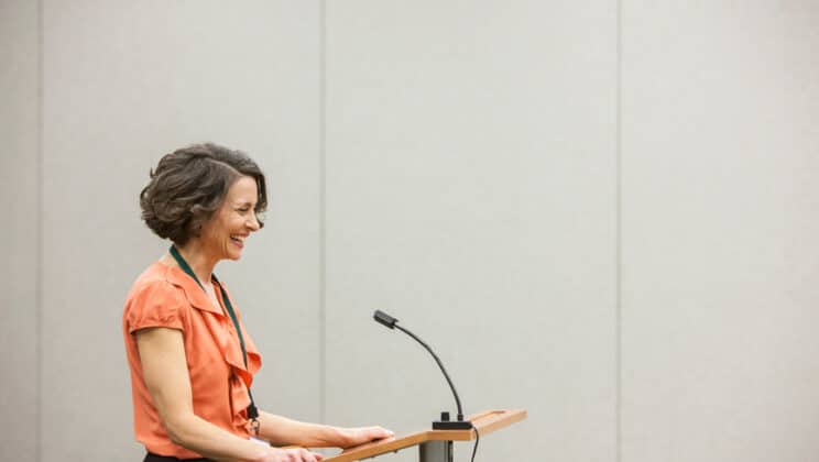 Caucasian businesswoman speaking from a podium to an audience in a conference room seminar setting.