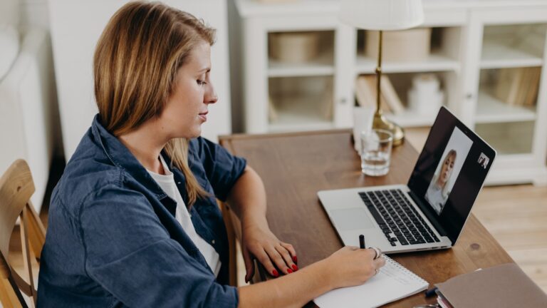 Side view of psychotherapist listening to client and writing in notepad during online psychotherapy session in modern office