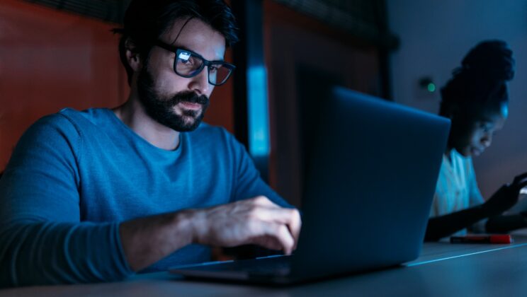 Side view of focused bearded male entrepreneur using netbook while working in dark office with screen hanging on wall near multiethnic coworkers