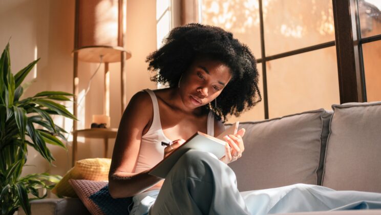 Young black woman at home, sitting on the couch and writing in her notebook.