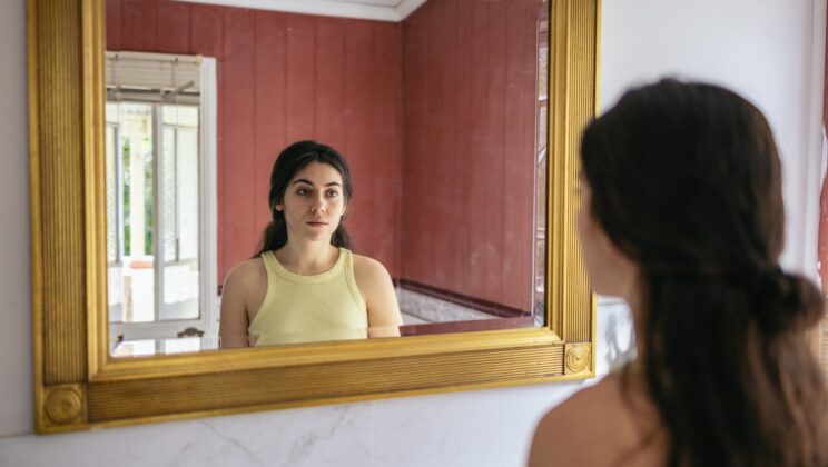 Young woman looking at herself in the mirror with sad expression in a red bathroom.