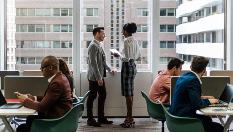 Side view of female secretary in formal wear standing and reading project to male manager in formal suit among busy office