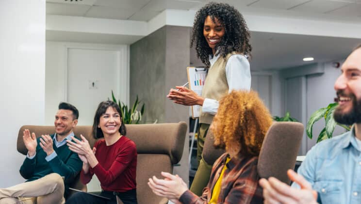 Diverse team clapping for a female colleague holding a clipboard in a bright business office setting.